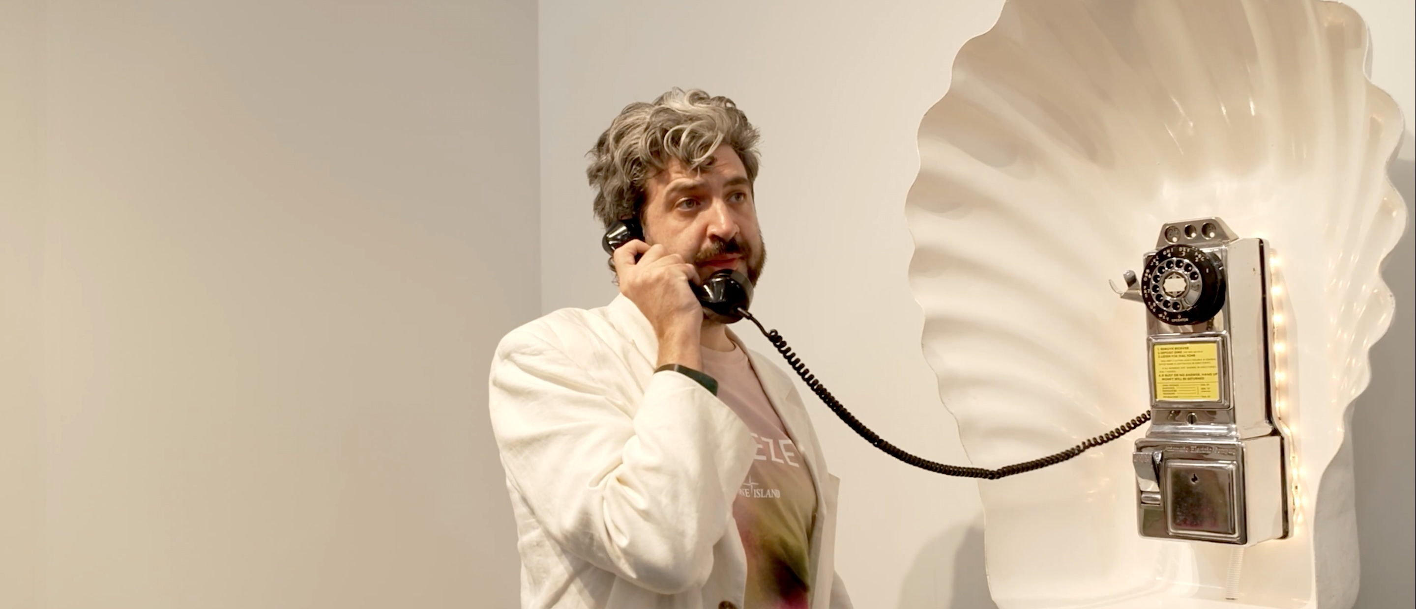 Matt McClean, Creative Director of Frieze Studios, speaks into a telephone mounted within a huge seashell on a gallery wall at Frieze Los Angeles 2025.